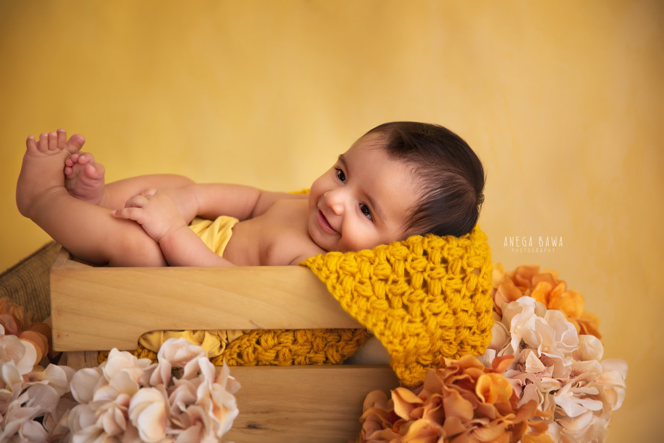 Captivating baby boy photoshoot featuring the delightful 6, 5, 4, 3-month-old against a cheerful yellow backdrop, nestled in a charming wooden cot adorned with vibrant orange and peach flowers. Skillfully captured by Anega Bawa, the esteemed baby photographer in Delhi, Gurgaon, and Noida.