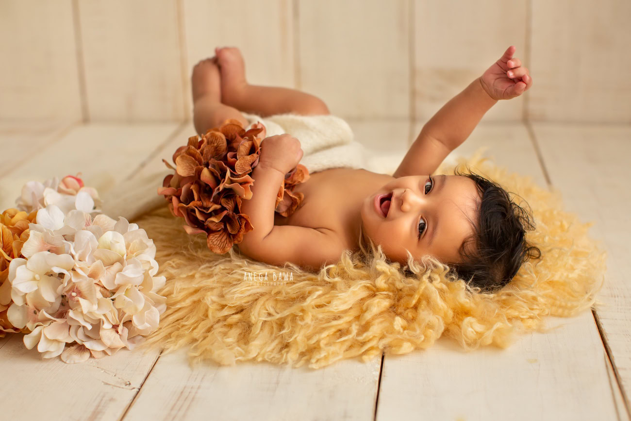 Captivating baby boy photoshoot featuring the delightful 6, 5, 4, 3-month-old on a vibrant yellow rug against a warm beige backdrop, surrounded by delicate peach and orange flowers. Skillfully captured by Anega Bawa, the esteemed baby photographer in Delhi, Gurgaon, and Noida.