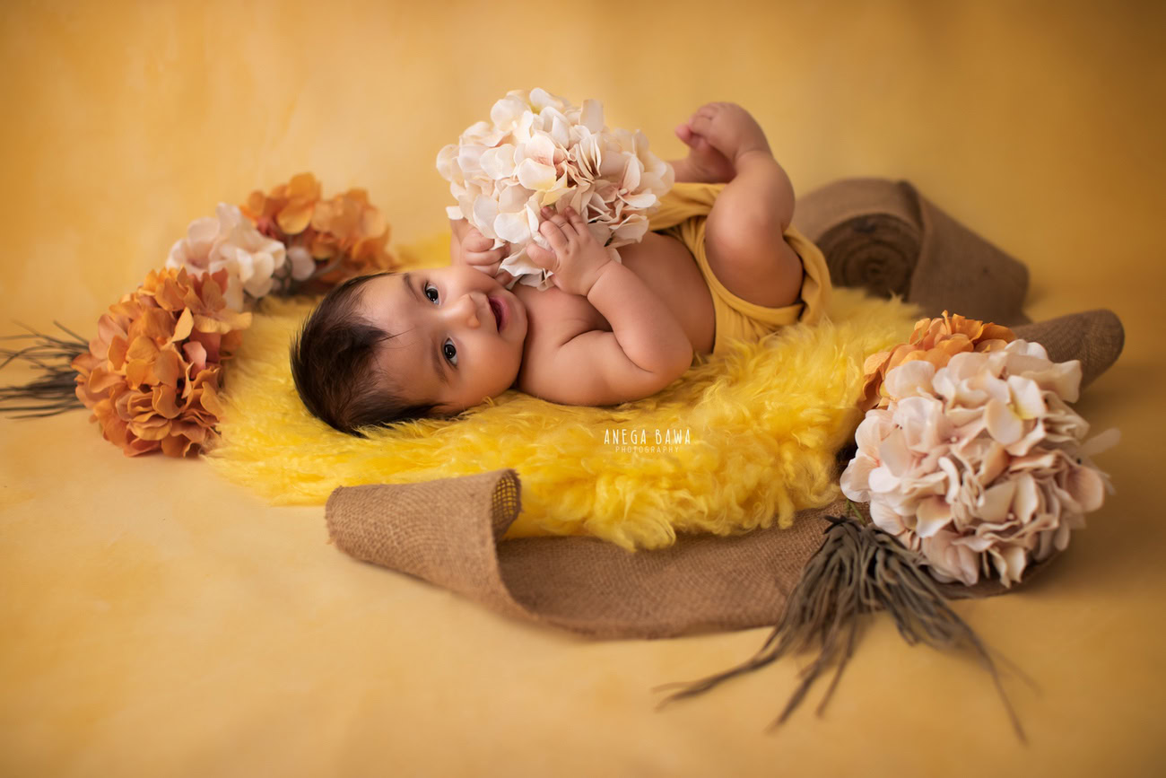 Captivating baby boy photoshoot featuring the delightful 6, 5, 4, 3-month-old on a vibrant yellow rug against a cheerful yellow backdrop, surrounded by vibrant orange and peach flowers. Skillfully captured by Anega Bawa, the esteemed baby photographer in Delhi, Gurgaon, and Noida.