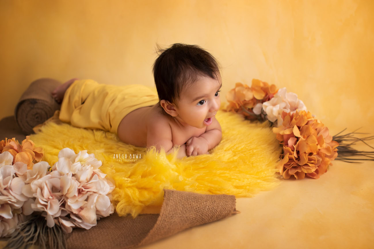 Captivating baby boy photoshoot showcasing the adorable 6, 5, 4, 3-month-old wrapped in a sunny yellow wrap, against a vibrant yellow backdrop, resting on a cozy yellow rug. Skillfully crafted by Anega Bawa, the premier baby photographer in Delhi, Gurgaon, and Noida.