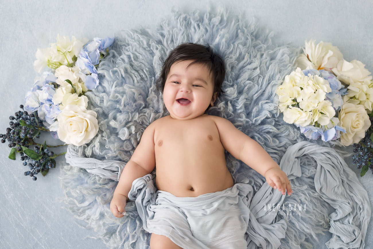 Capture the joy of your baby boy at 3, 4, 5, or 6 months with a delightful photoshoot featuring a cozy grey rug, set against a neutral grey backdrop, accented with white flowers. Witness those precious laughing moments frozen in time. Reach out to Anega Bawa for unforgettable baby boy photoshoots in Delhi, Gurgaon, or Noida.