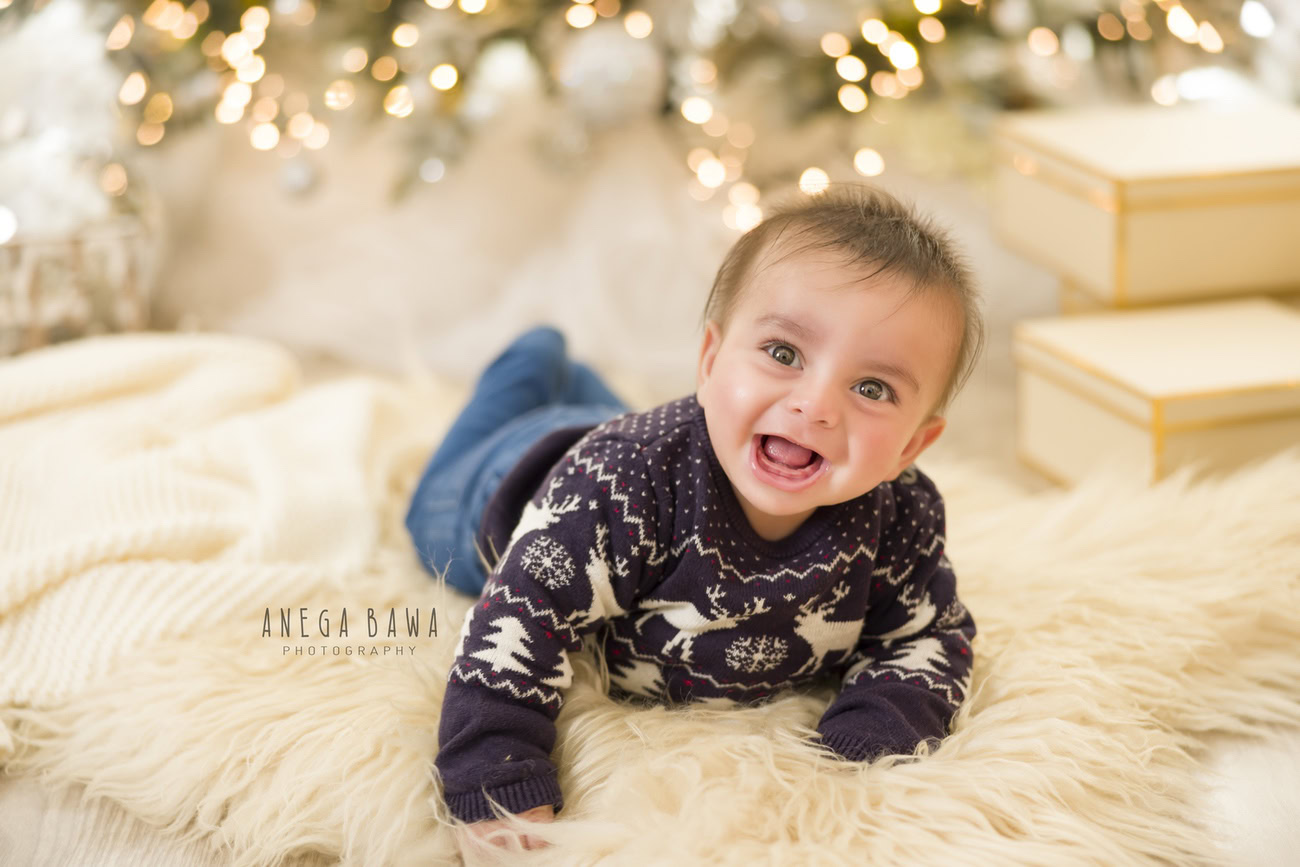 Capture the holiday spirit with your little one aged 3, 4, 5, or 6 months in an adorable Xmas jumper, flashing a heartwarming smile, set against a cozy beige rug adorned with golden lights. Contact Anega Bawa for delightful baby boy photoshoots in Delhi, Gurgaon, or Noida.