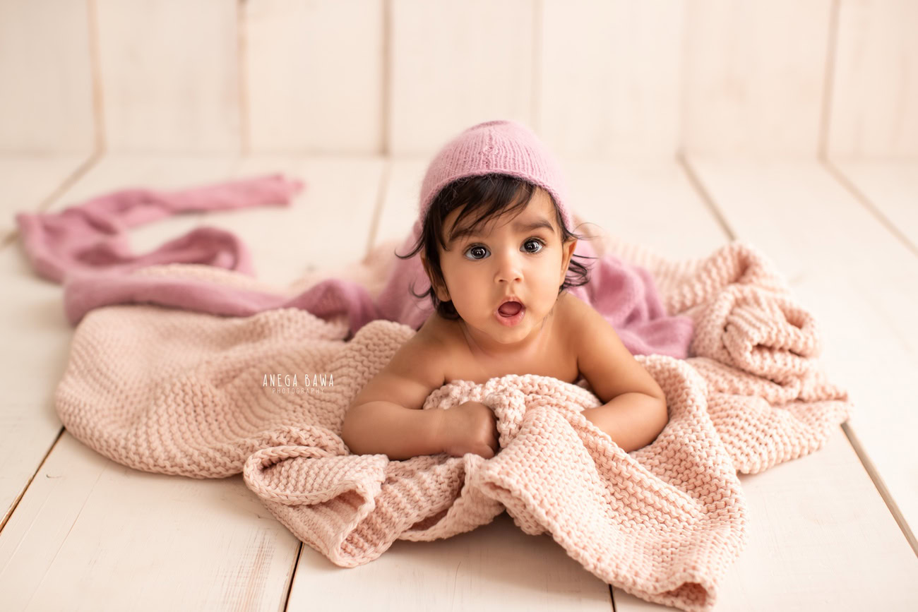 Captivating baby boy photoshoot ideas featuring the precious 5, 4, 3, 6-month-old against a warm beige backdrop, adorned with a soft pink sheet, striking a cute pose. Skillfully crafted by Anega Bawa, the premier baby photographer in Delhi, Gurgaon, and Noida.
