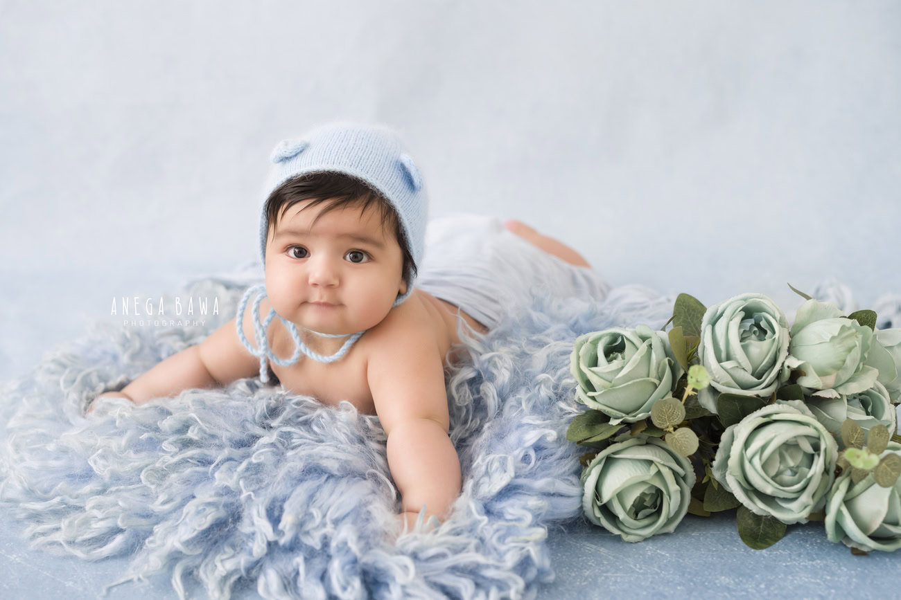 Create precious memories of your little boy aged 3, 4, 5, or 6 months with a charming blue rug, set against a serene blue backdrop, adorned with a cute cap and surrounded by green flowers. Contact Anega Bawa for captivating baby boy photoshoots in Delhi, Gurgaon, or Noida.