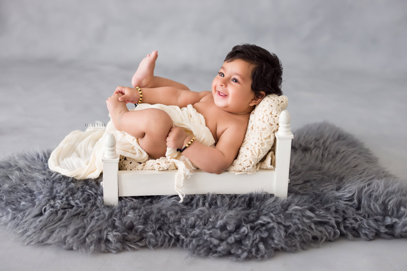 Baby boy photoshoot with grey rug, grey backdrop, white wooden cot, and white wrap for babies aged 5, 4, 3, or 6 months by Anega Bawa in Delhi, Gurgaon, Noida.