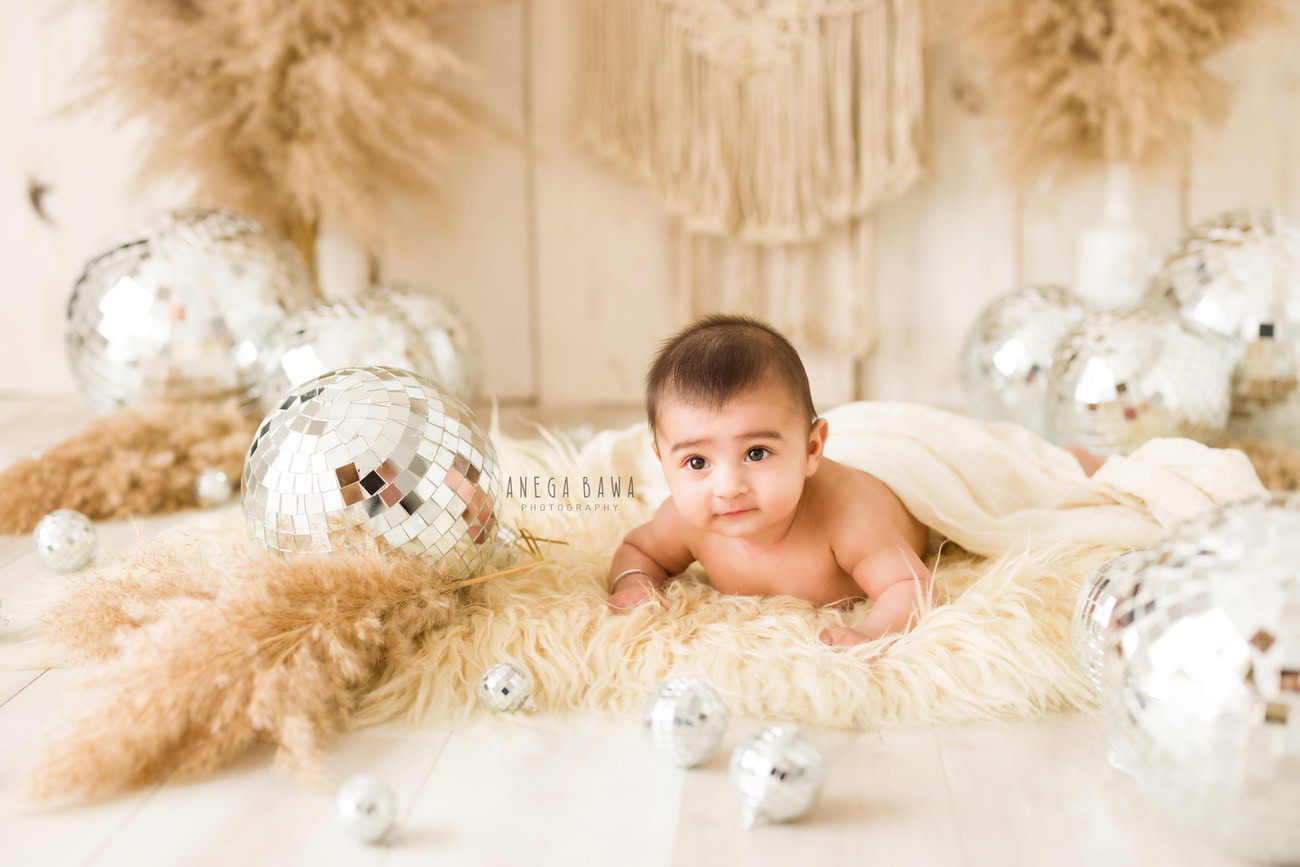 Capture the precious moments of your little princess at 3, 4, 5, or 6 months old, adorned in a delightful beige rug, surrounded by shimmering discoballs, lying on an off-white sheet against a serene beige backdrop. Contact Anega Bawa for enchanting baby girl photoshoots in Delhi, Gurgaon, or Noida.