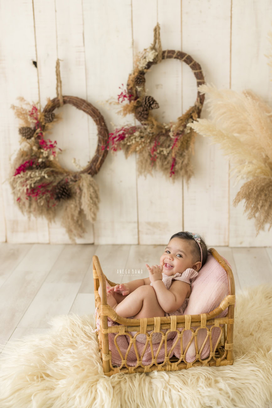 Capture the charm of a baby girl at 6, 5, 3, or 4 months with a wooden cot adorned with a beige rug, set against a backdrop of wooden frames and shrubs on a beige backdrop. Photographed by Anega Bawa in Delhi, Gurgaon, Noida.