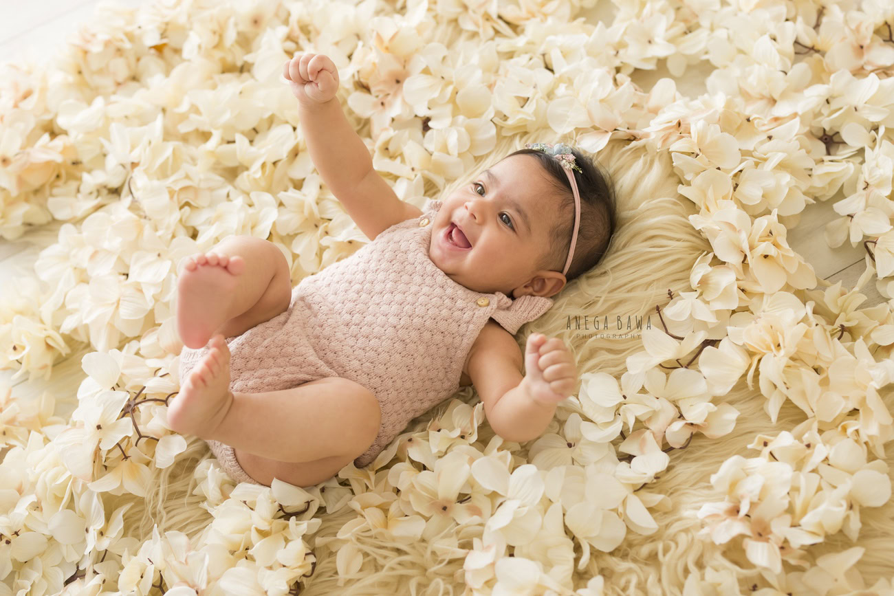 Embrace the sweetness of a baby girl aged 6, 5, 4, or 3 months in a baby pink bodysuit, complemented by a beige rug and an adorable headband, captured in a joyful smiling pose. Photography by Anega Bawa in Delhi, Gurgaon, Noida.
