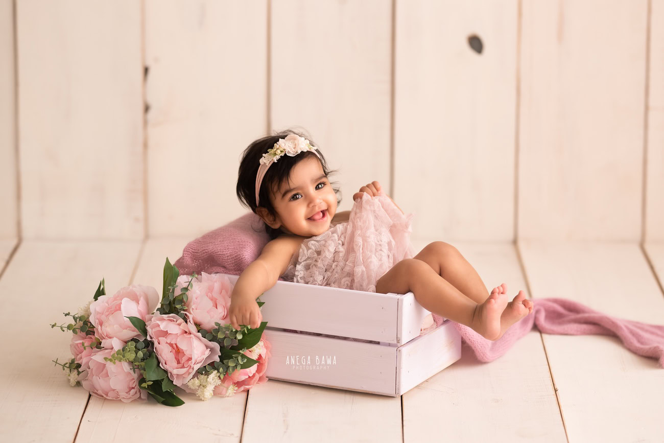 Captivating baby girl photoshoot showcasing the charming 6, 5, 4, 3-month-old against a warm beige backdrop, wrapped in a delicate pink net adorned with pink flowers, striking a cute smiling pose. Skillfully crafted by Anega Bawa, the premier baby photographer in Delhi, Gurgaon, and Noida.