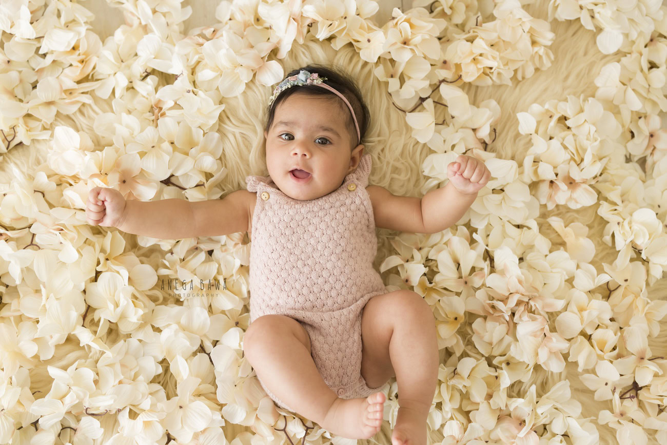 A baby girl, aged 6, 5, 4, or 3 months, posing in a baby pink bodysuit on a beige rug surrounded by white flowers on the floor, against a beige backdrop, perfect for a baby photoshoot in Delhi, Gurgaon, or Noida with Anega Bawa.