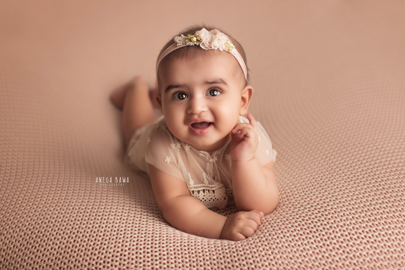 Captivating baby girl photoshoot showcasing the adorable 6, 5, 4, 3-month-old in a cute headband, striking a smiling pose against a rich brown backdrop. Skillfully crafted by Anega Bawa, the premier baby photographer in Delhi, Gurgaon, and Noida.