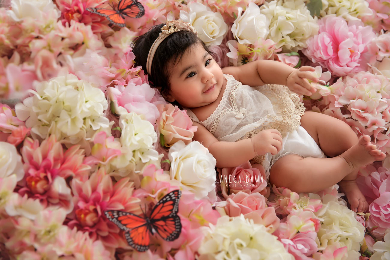 Captivating baby girl photoshoot featuring the delightful 6, 5, 4, 3-month-old adorned with a cute headband, surrounded by multicolour flowers and a butterfly, against a charming floral backdrop. Skillfully crafted by Anega Bawa, the premier baby photographer in Delhi, Gurgaon, and Noida.