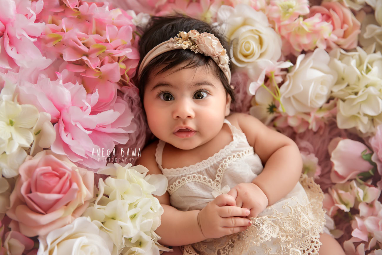 Captivating baby girl photoshoot showcasing the delightful 6, 5, 4, 3-month-old adorned with a cute headband, surrounded by pink and white flowers. Skillfully crafted by Anega Bawa, the premier baby photographer in Delhi, Gurgaon, and Noida.