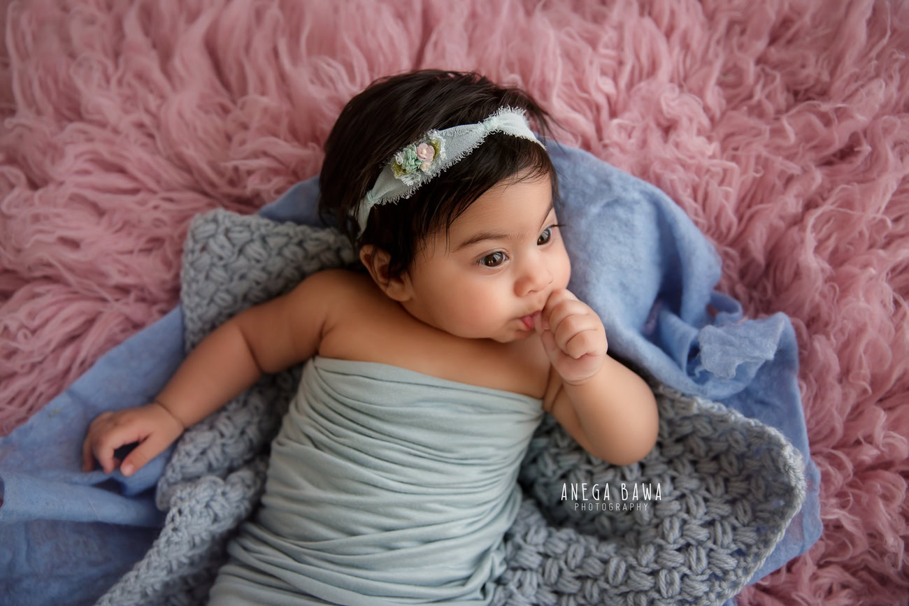 Captivating baby girl photoshoot featuring the charming 6, 5, 4, 3-month-old on a soft pink rug, wrapped in a delicate blue wrap, adorned with a cute headband, against a lovely pink backdrop. Skillfully crafted by Anega Bawa, the premier baby photographer in Delhi, Gurgaon, and Noida.