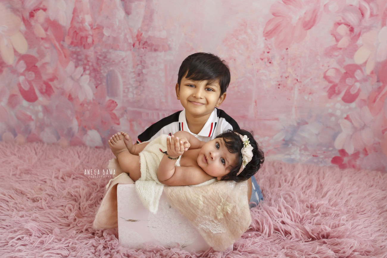 Captivating baby girl photoshoot featuring the endearing 6, 5, 4, 3-month-old on a soft pink rug, accompanied by a sibling, against a matching pink backdrop. Skillfully crafted by Anega Bawa, the premier baby photographer in Delhi, Gurgaon, and Noida.
