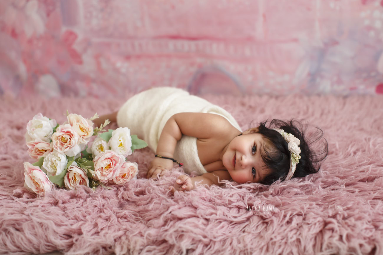 Captivating baby girl photoshoot presenting the adorable 6, 5, 4, 3-month-old on a soft pink rug, wrapped in a pristine white wrap, surrounded by charming pink and white flowers, against a lovely pink backdrop. Skillfully crafted by Anega Bawa, the premier baby photographer in Delhi, Gurgaon, and Noida.
