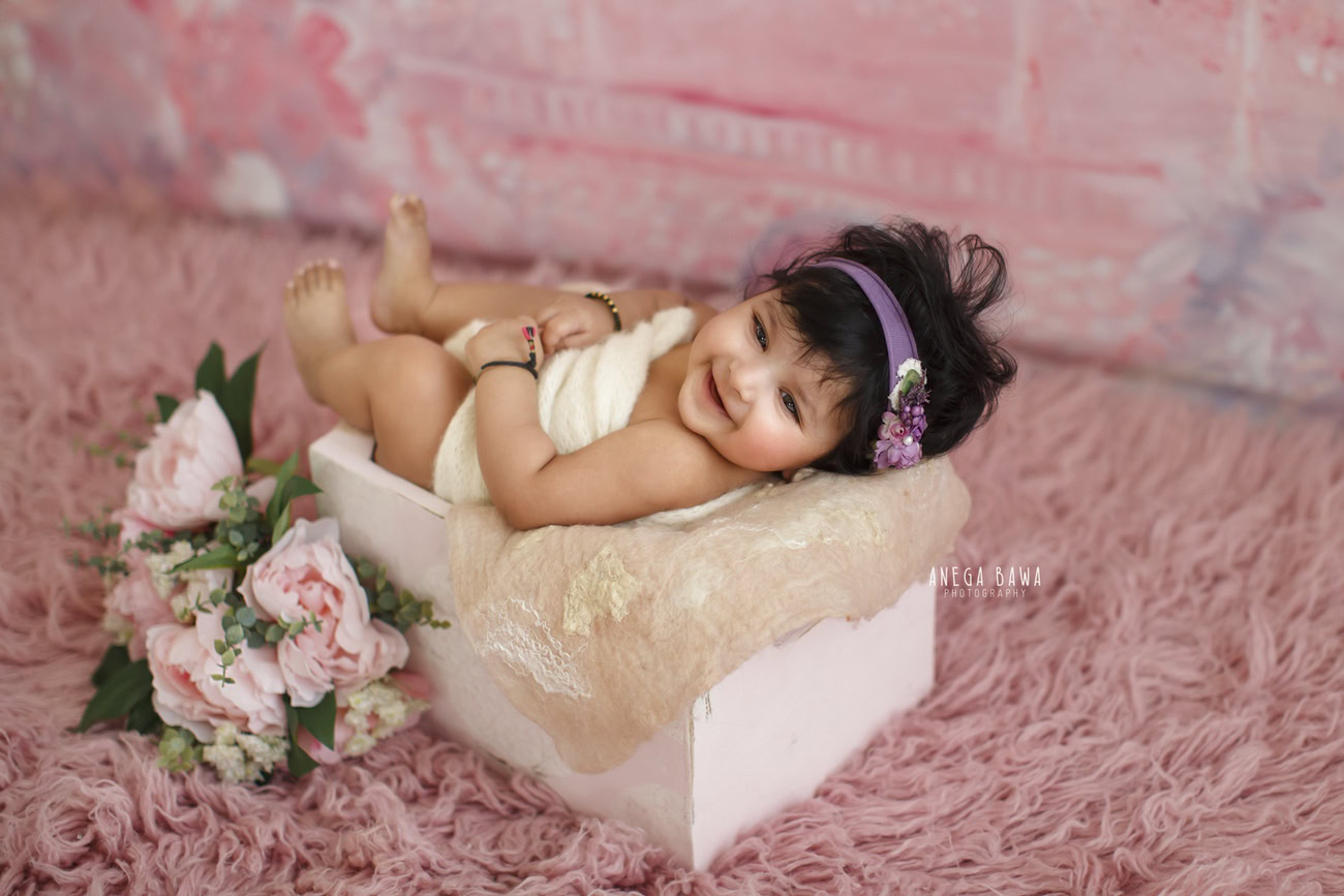 Captivating baby girl photoshoot showcasing the delightful 6, 5, 4, 3-month-old in a white wooden basket, wrapped in a pristine white wrap, adorned with flowers and a cute headband, against a charming pink backdrop. Skillfully crafted by Anega Bawa, the premier baby photographer in Delhi, Gurgaon, and Noida.