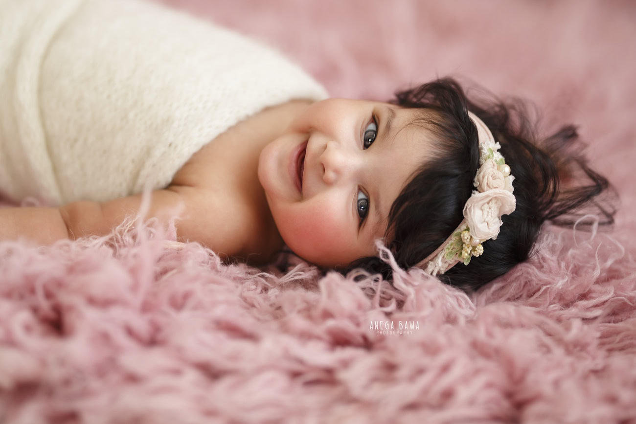 Captivating baby girl photoshoot featuring the adorable 6, 5, 4, 3-month-old wrapped in a pristine white wrap adorned with a cute headband, against a charming pink backdrop. Skillfully crafted by Anega Bawa, the premier baby photographer in Delhi, Gurgaon, and Noida.