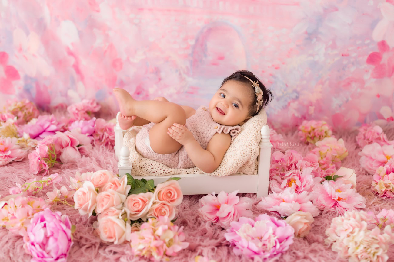 A baby girl photographer captures adorable moments of infants aged 3, 4, 5, or 6 months against a charming pink backdrop adorned with pink and peach flowers. The ideal setting for a baby photoshoot in Delhi, Gurgaon, or Noida with Anega Bawa.