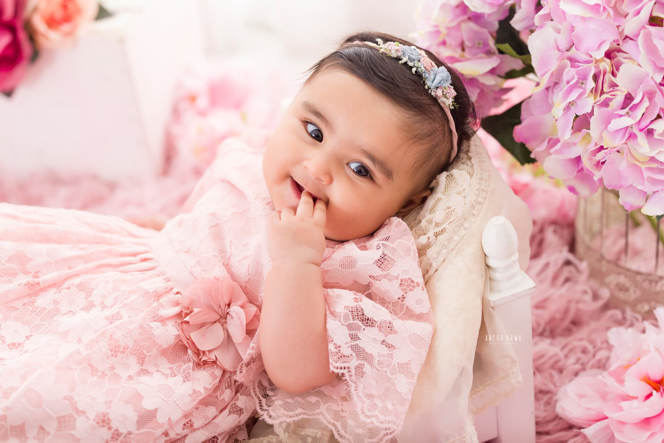A baby girl photographer captures the charm of infants aged 3, 4, 5, or 6 months against a backdrop adorned with pink flowers, accentuated with a cute headband. Ideal for a baby photoshoot in Delhi, Gurgaon, or Noida with Anega Bawa.