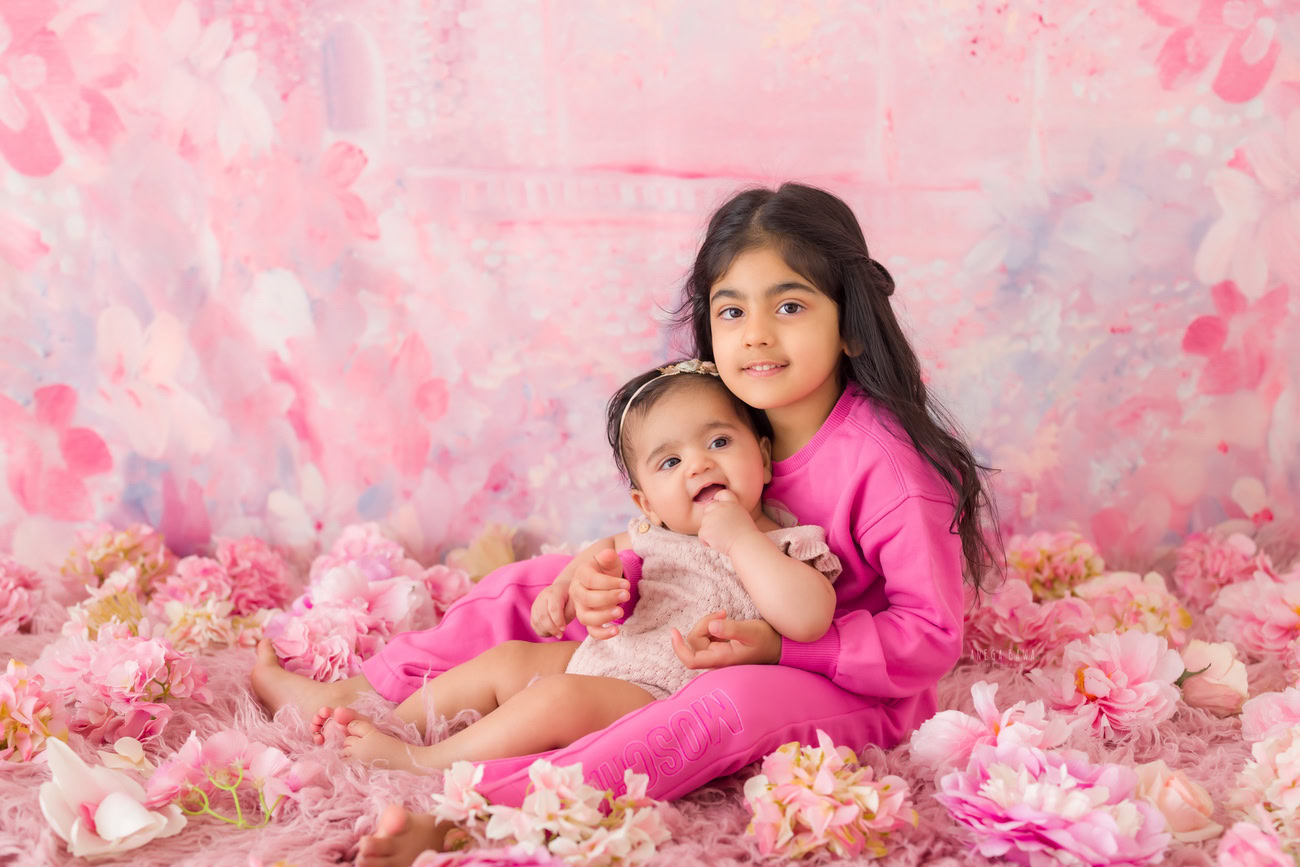 Capture the adorable bond between siblings, aged 3, 4, 5, or 6 months, amidst a backdrop adorned with pink flowers. Perfect for a baby photoshoot in Delhi, Gurgaon, or Noida with Anega Bawa.