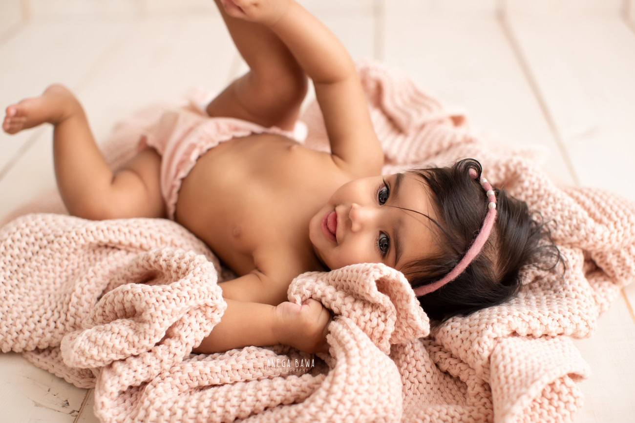 Captivating baby girl photoshoot by Anega Bawa, the esteemed baby photographer in Delhi, Gurgaon, and Noida, featuring the precious 3, 4, 5, 6-month-old in a cute lying pose on a soft pink sheet.