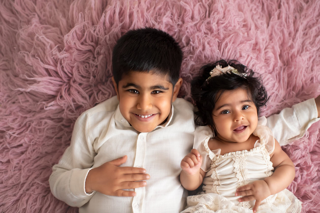 Captivating baby girl photoshoot by Anega Bawa, the esteemed baby photographer in Delhi, Gurgaon, and Noida, featuring the adorable 3, 4, 5, 6-month-old posing cutely with a sibling on a soft pink rug, adorned in a white dress.