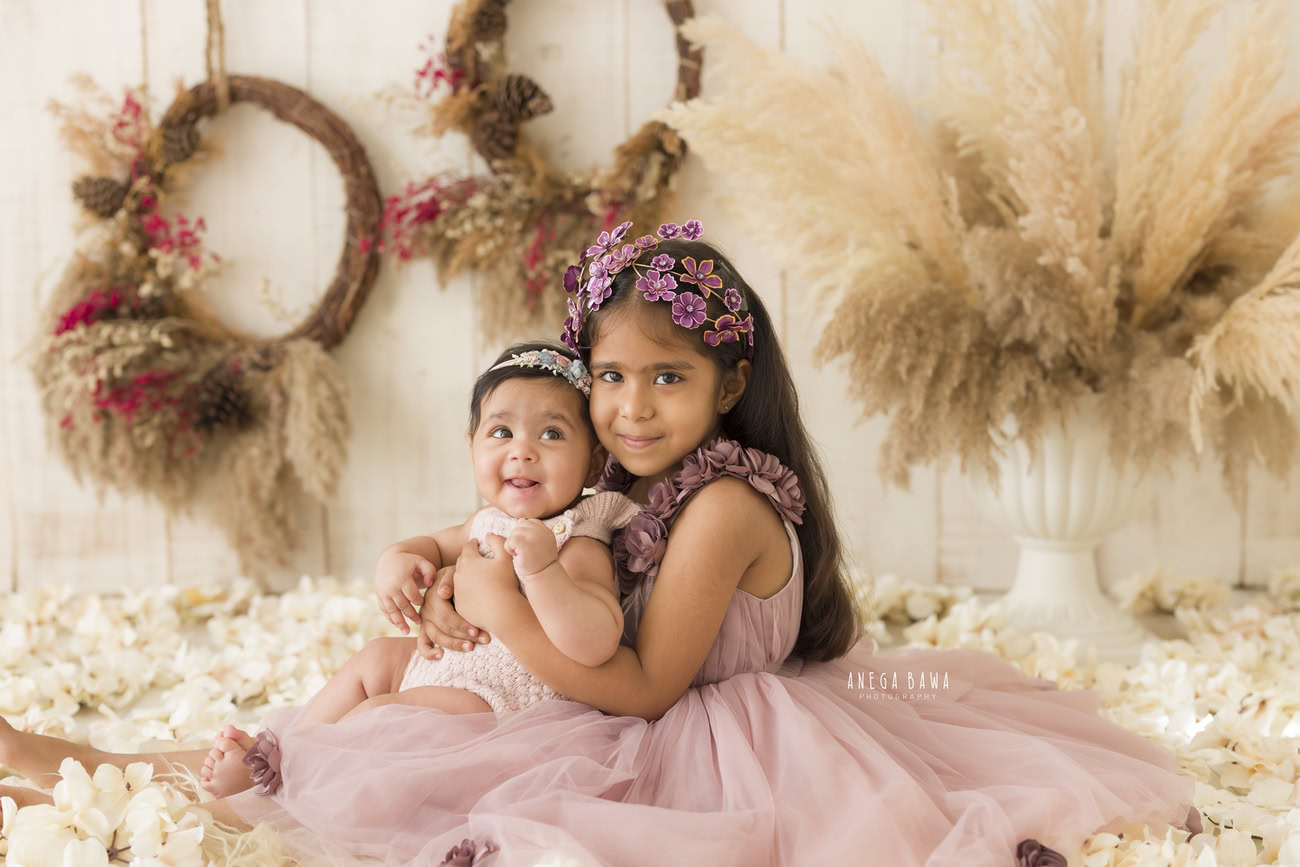Immerse your little angel in a picturesque scene with a beige backdrop adorned with wooden floral frames and surrounded by brown shrubs. For sibling moments captured beautifully against a matching beige backdrop, book your baby girl's photography session in Delhi, Gurgaon, or Noida with Anega Bawa today!