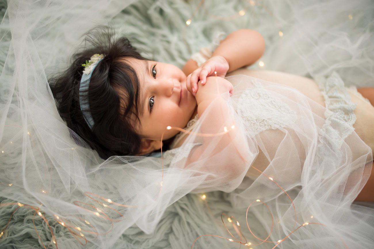 Capture the innocence of your 3 to 5-month-old baby girl in a charming photoshoot. Picture her in a grey net dress, resting on a soft grey rug, illuminated by golden lights against a serene grey backdrop. Contact Anega Bawa for exquisite baby girl photography in Delhi, Gurgaon, or Noida.