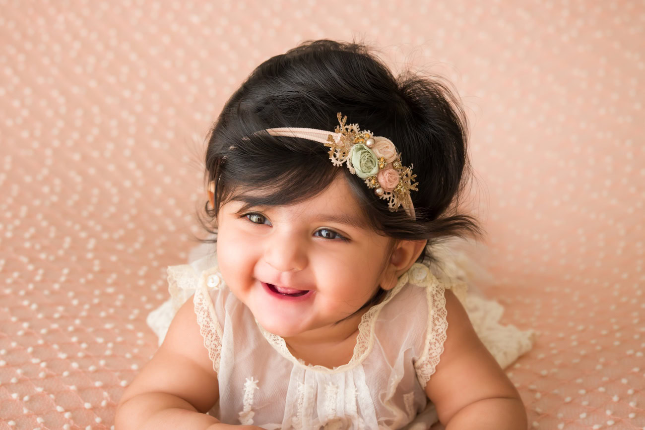 Create lasting memories of your 3 to 5-month-old baby girl with a charming peach backdrop, adorned with a cute headband, captured in a smiling pose. Contact Anega Bawa for professional baby girl photography in Delhi, Gurgaon, or Noida.