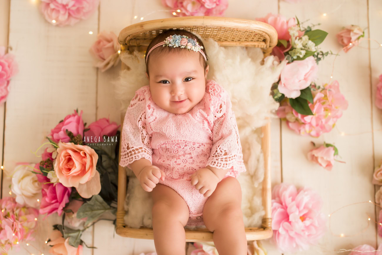 Captivating baby girl photography by Anega Bawa, the renowned baby photographer in Delhi, Gurgaon, and Noida, showcasing the adorable 6, 4, 3, 5-month-old in a charming pink babysuit and a cute headband, surrounded by pink flowers against a warm beige backdrop.