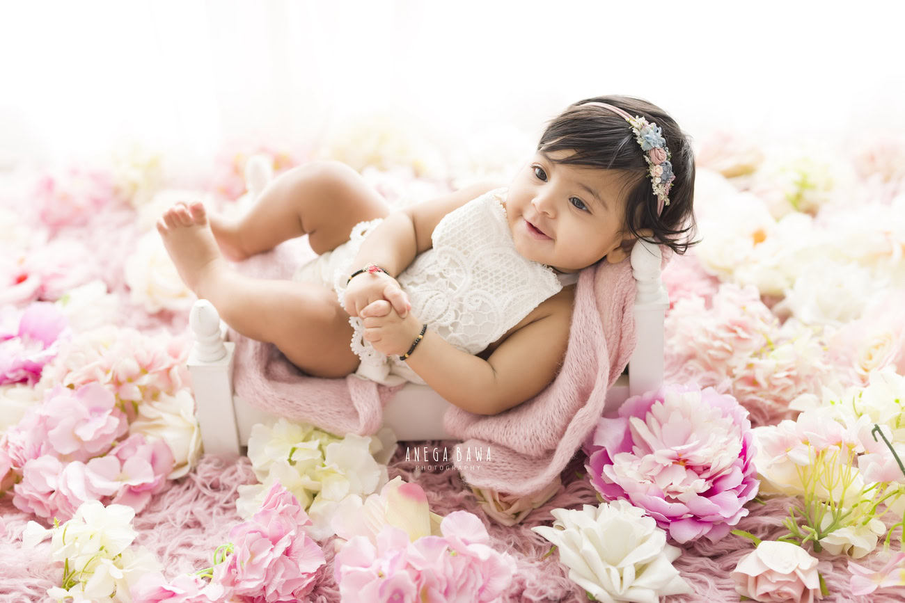 Adorn your little one in a cute headband amidst pink flowers, set against a pink sheet and a white backdrop for a charming photoshoot with Anega Bawa in Delhi, Gurgaon, or Noida.