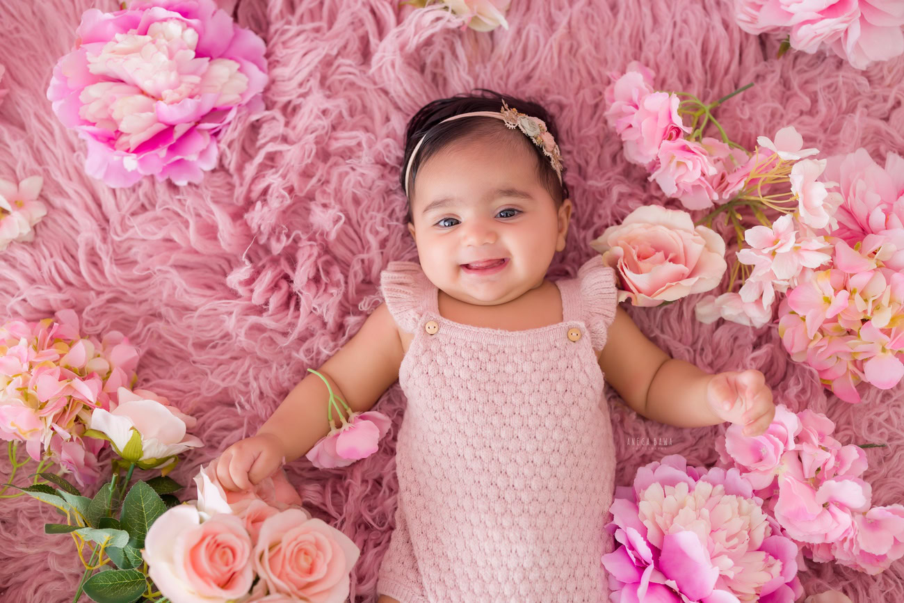 Capture adorable moments with your baby girl on a pink rug surrounded by flowers against a pink backdrop. Book a photoshoot with Anega Bawa in Delhi, Gurgaon, or Noida.