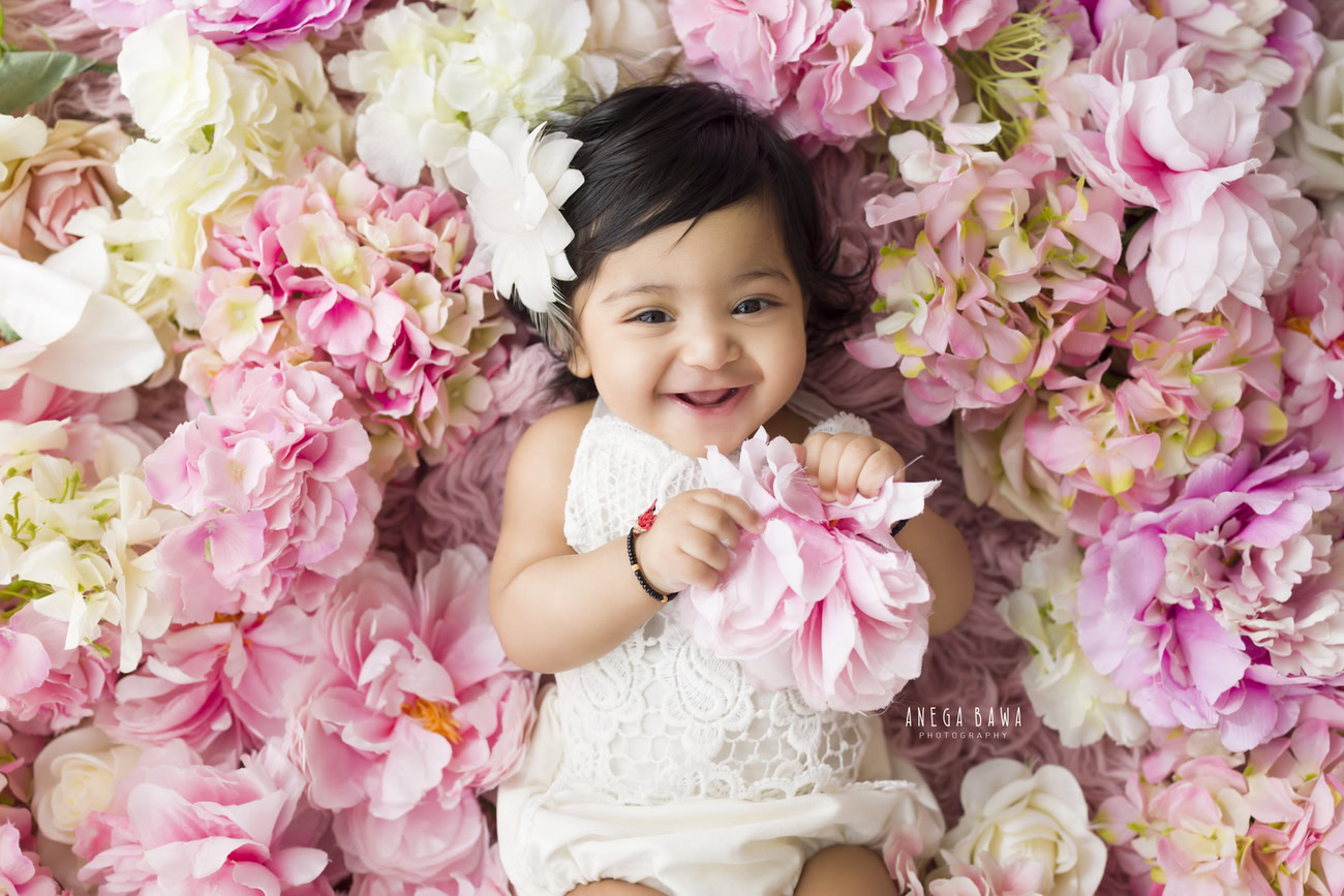 Capture the innocence of your little one with a cute smiling pose, adorned with white flowers in her hair, against a floral backdrop. Book a session with Anega Bawa for baby girl photography in Delhi, Gurgaon, or Noida.