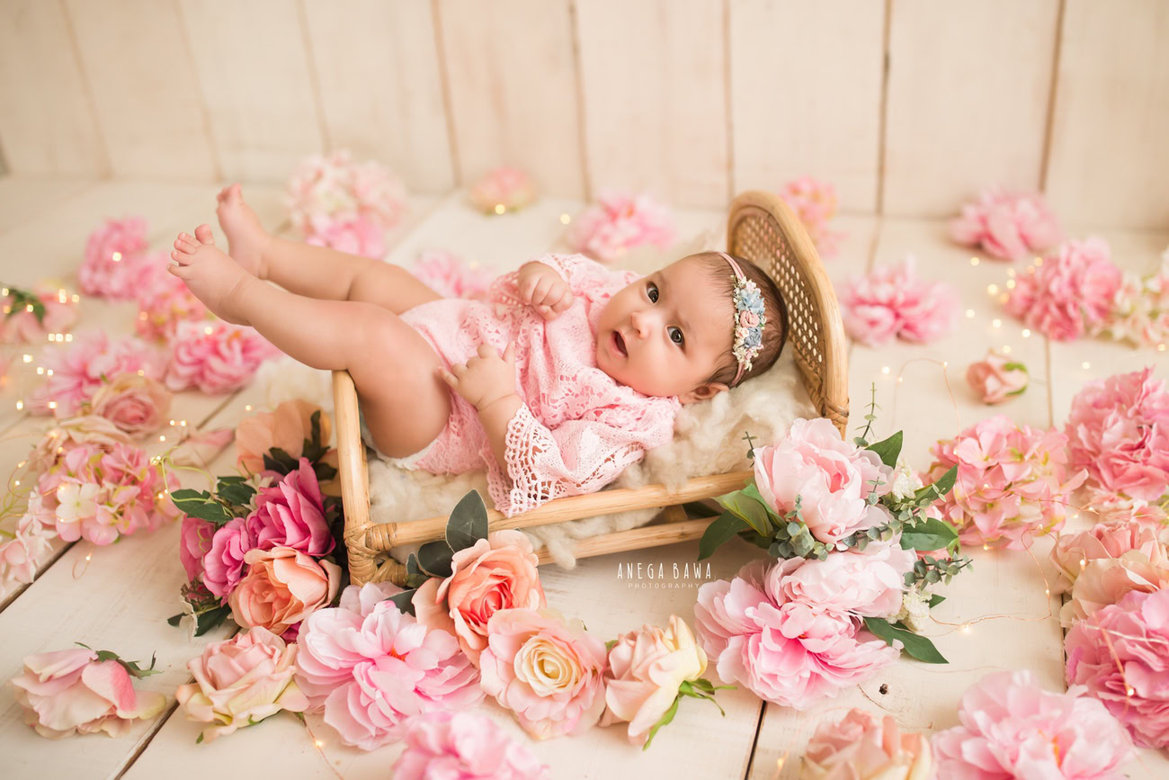 Capture precious moments of your 3 to 5-month-old baby girl in a charming wooden cot, dressed in an adorable pink baby suit, adorned with a pink flowers headband. Book your session with Anega Bawa in Delhi, Gurgaon, or Noida.