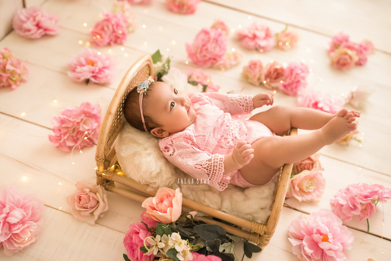 Captivating baby girl photography by Anega Bawa, the renowned baby photographer in Delhi, Gurgaon, and Noida, presenting the delightful 6, 4, 3, 5-month-old in a wooden cot adorned in a pink babysuit, with pink flowers scattered on the floor, against a warm beige backdrop.