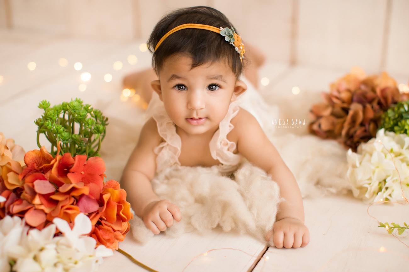 Celebrate your baby girl's milestones with a photoshoot featuring a beige backdrop, complemented by golden lights and accents of brown and orange flowers. Contact Anega Bawa for stunning captures in Delhi, Gurgaon, or Noida.