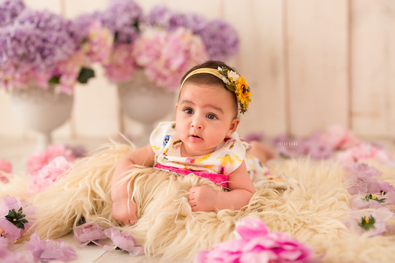 Capture the innocence of your baby girl aged 3, 4, 5, or 6 months with a delightful photoshoot featuring a beige rug, a pink lavender flower vase, and a beige backdrop. Contact Anega Bawa for a session in Delhi, Gurgaon, or Noida.