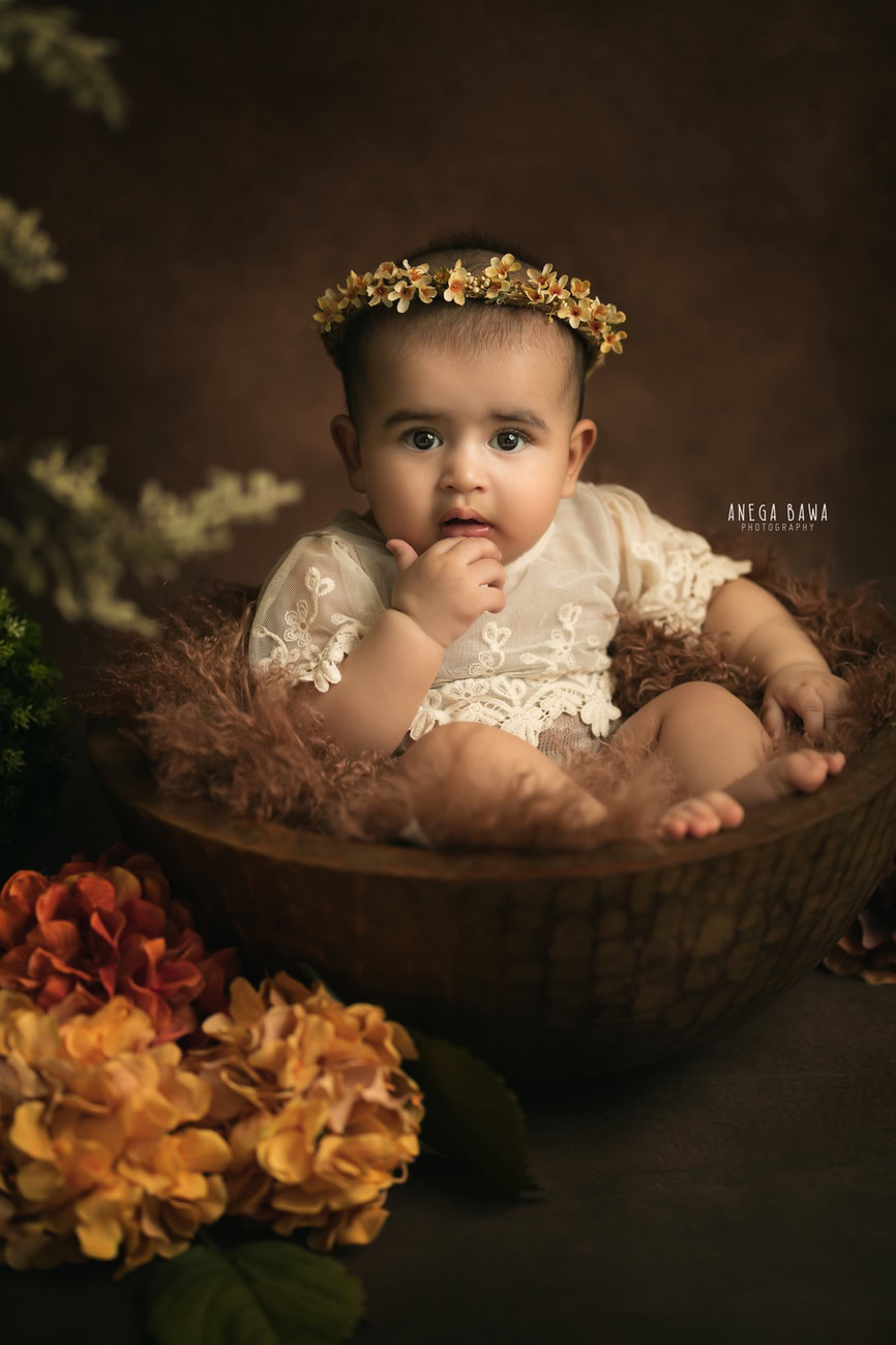 Captivating baby girl photoshoot by Anega Bawa, the esteemed baby photographer in Delhi, Gurgaon, and Noida, featuring the adorable 3, 4, 5, 6-month-old in a cute pose, nestled in a brown basket against a rich brown backdrop.