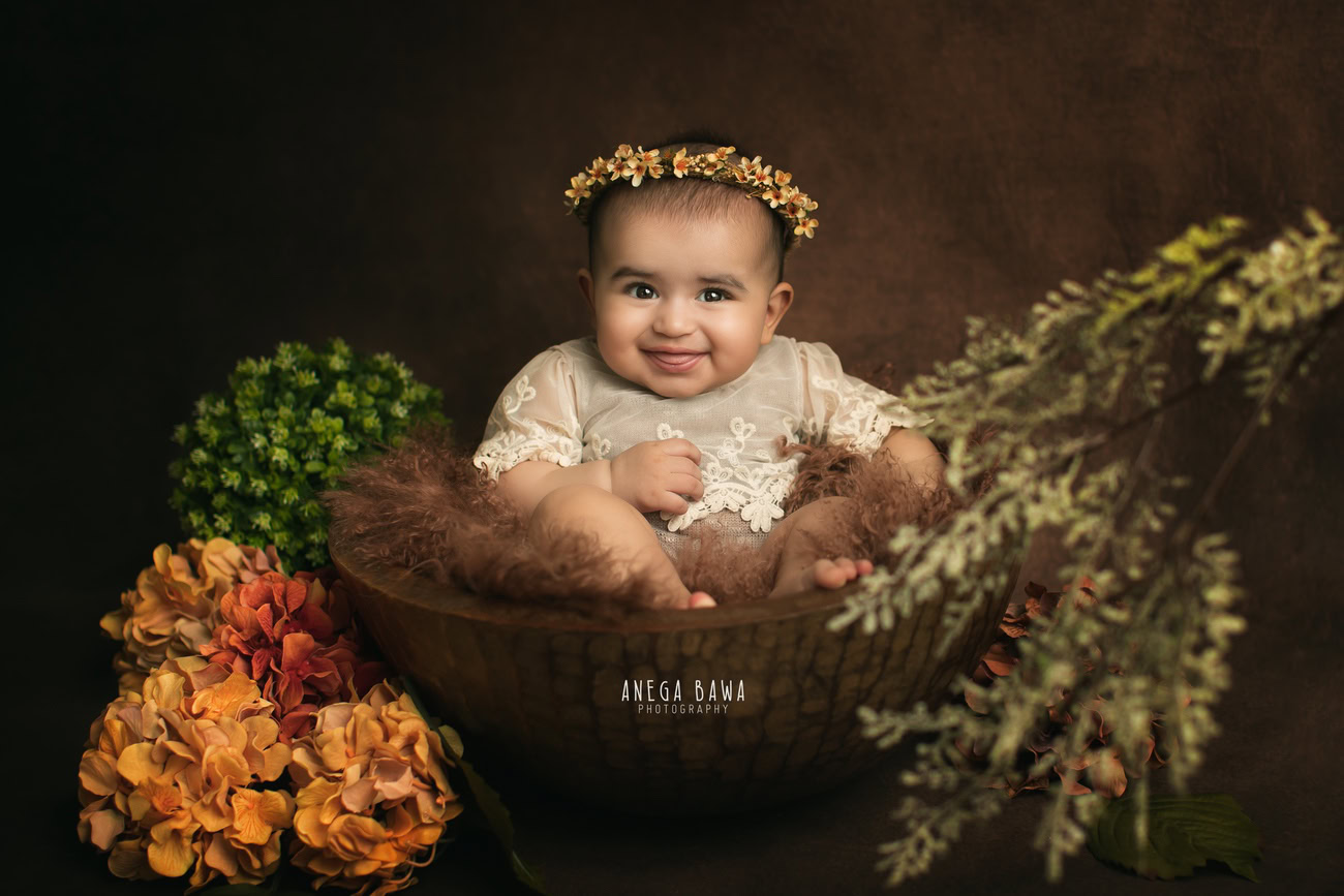 Captivating baby girl photoshoot by Anega Bawa, the esteemed baby photographer in Delhi, Gurgaon, and Noida, featuring the precious 3, 4, 5, 6-month-old adorned with a floral headband, nestled in a brown basket against a rich brown backdrop.