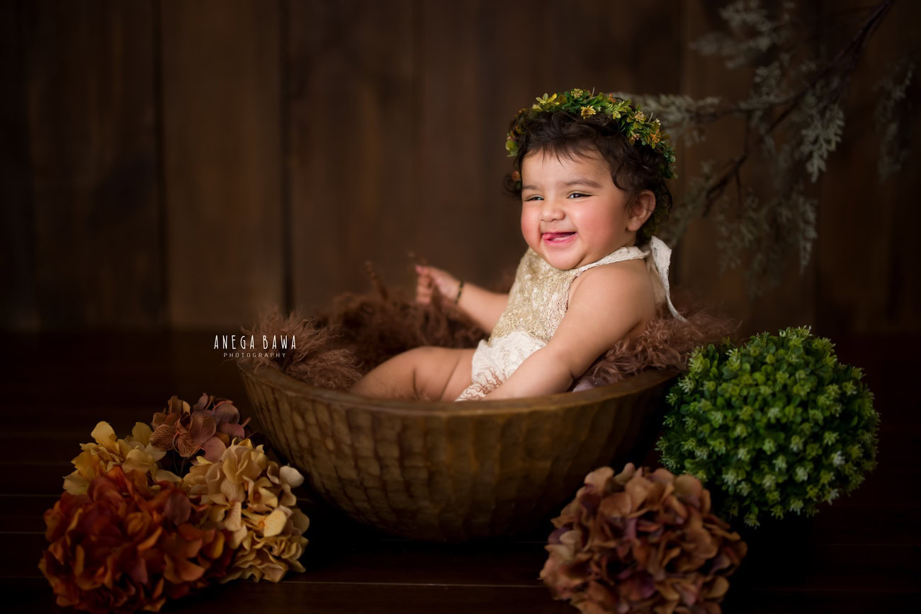 Immerse in the enchanting beauty of your little one with a brown basket surrounded by vibrant orange and green flowers, capturing a delightful seating pose against a brown backdrop. Contact Anega Bawa for an unforgettable photoshoot experience in Delhi, Gurgaon, or Noida.