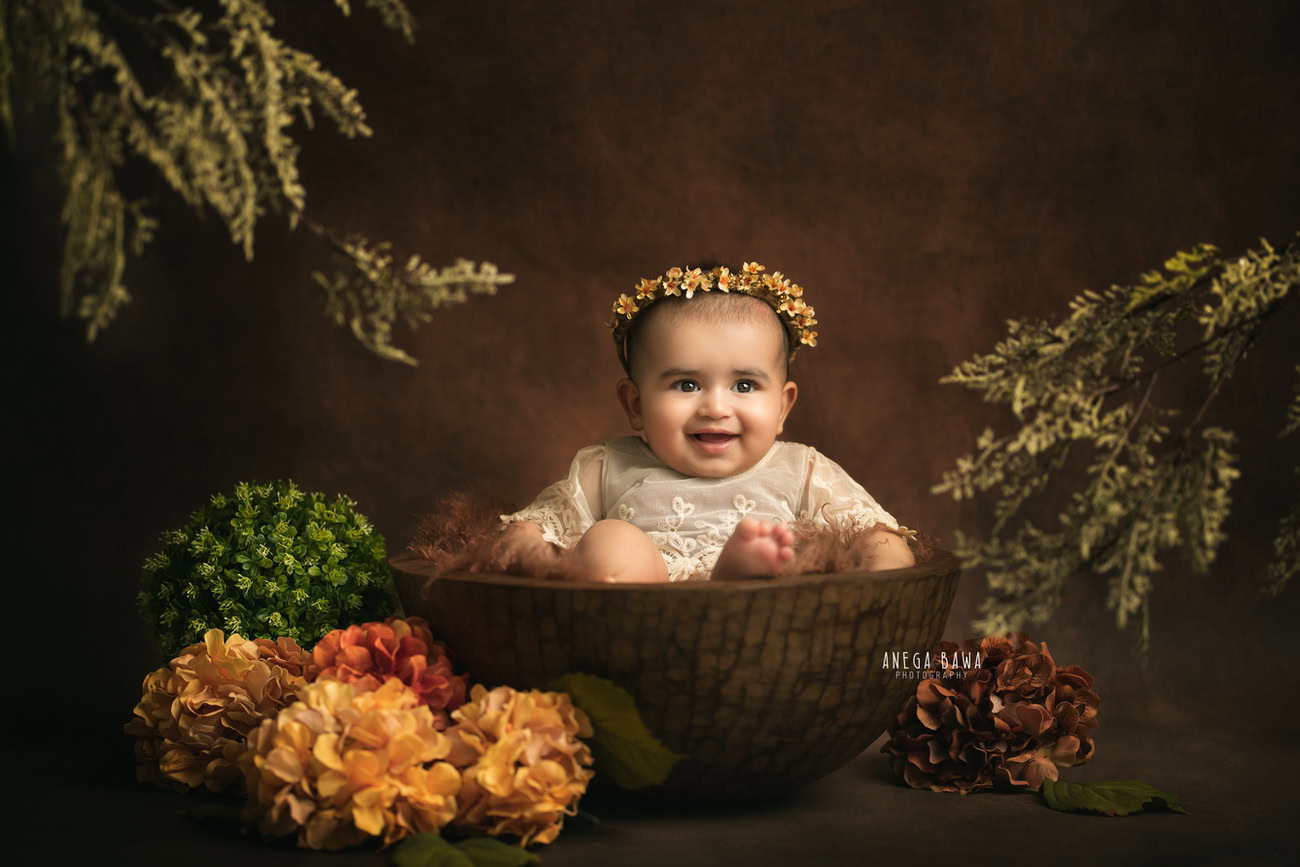 Captivating baby girl photoshoot by Anega Bawa, the esteemed baby photographer in Delhi, Gurgaon, and Noida, showcasing the precious 3, 4, 5, 6-month-old adorned with a floral headband, nestled in a brown basket surrounded by yellow and orange flowers against a rich brown backdrop.