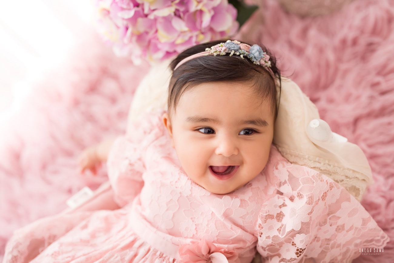 Capture precious moments of your baby girl aged 3, 4, 5, or 6 months in a delightful pink dress, posed on a soft pink rug with a charming headband, against a pristine white backdrop. Book your session with Anega Bawa in Delhi, Gurgaon, or Noida.