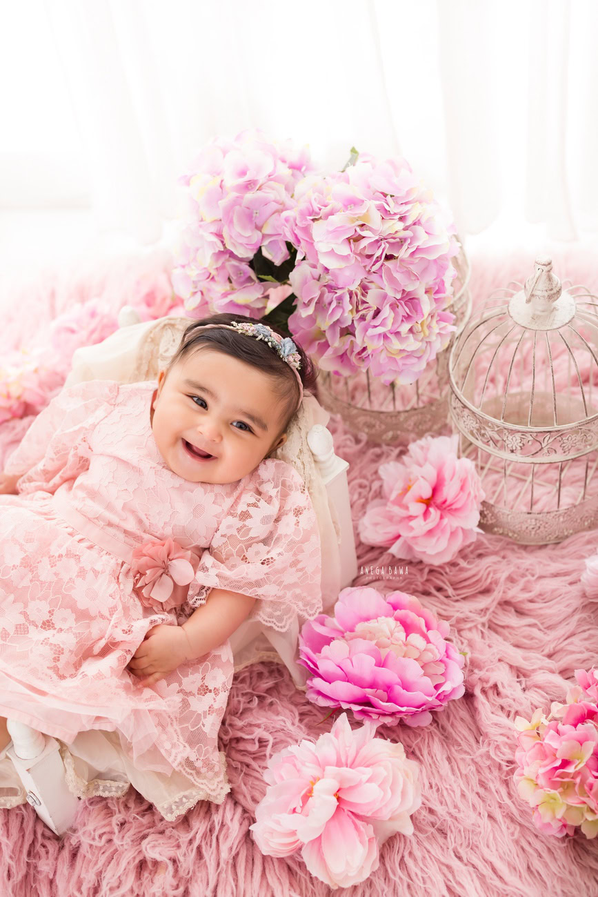Document the charm of your baby girl aged 3, 4, 5, or 6 months in a delightful pink dress, resting on a soft pink rug, against a pristine white backdrop adorned with pink flowers. Book your session with Anega Bawa in Delhi, Gurgaon, or Noida.