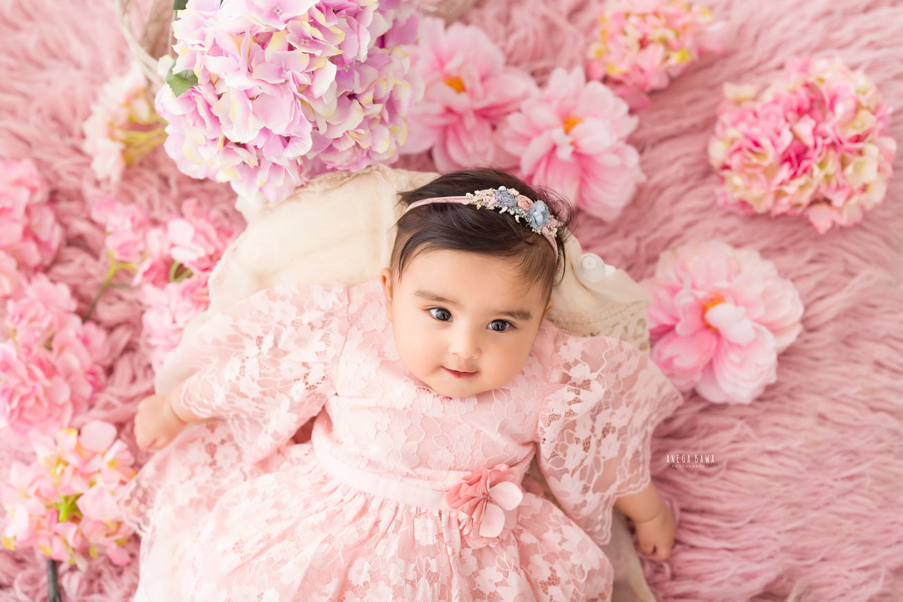 Capture the innocence and sweetness of your baby girl aged 3, 4, 5, or 6 months in a charming pink dress, adorned with a cute headband, resting on a soft pink rug. Contact Anega Bawa for your baby girl's photoshoot in Delhi, Gurgaon, or Noida.