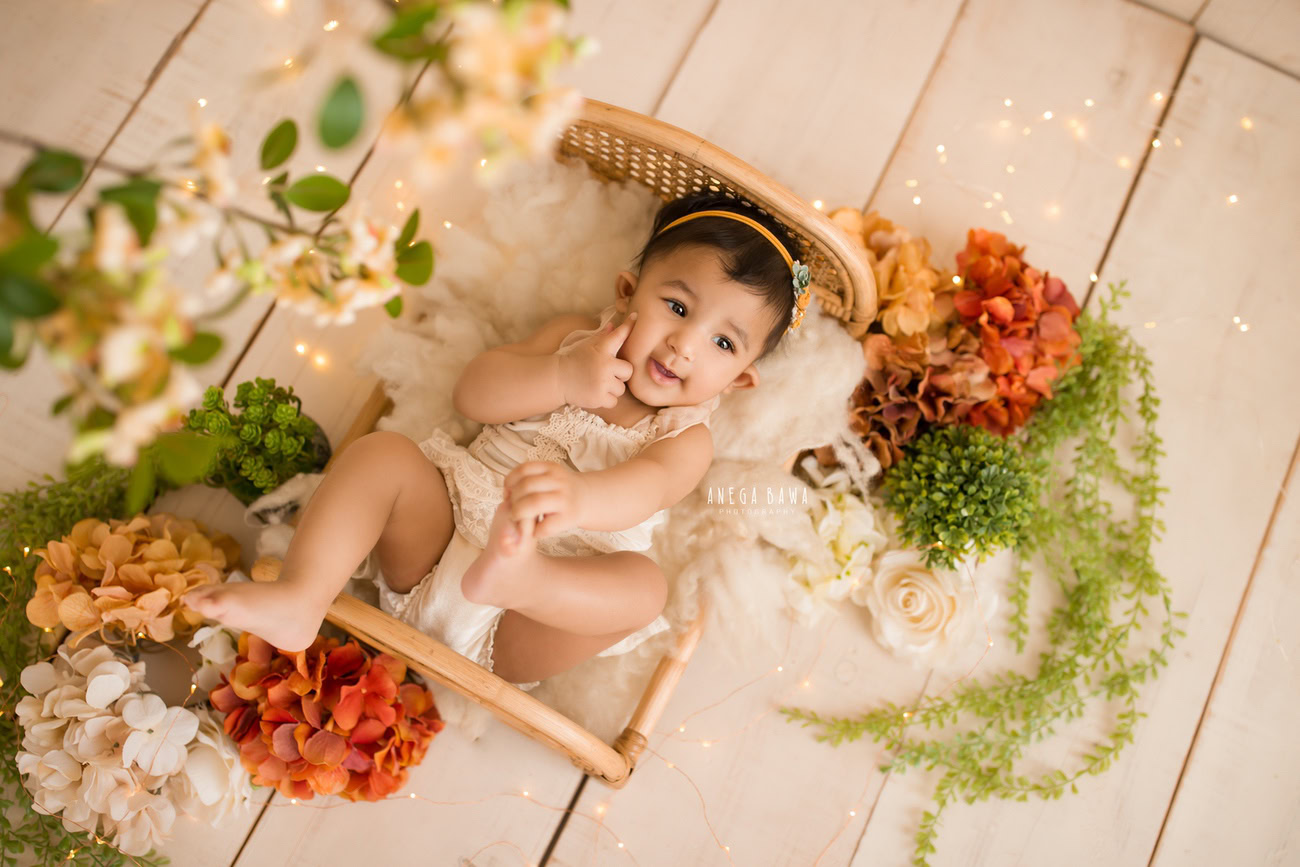 Capture the charm of your baby girl's milestones with a captivating photoshoot! Featuring a wooden cot adorned with brown and orange flowers against a soothing beige backdrop, this session promises timeless memories. Contact Anega Bawa to book your session in Delhi, Gurgaon, or Noida.