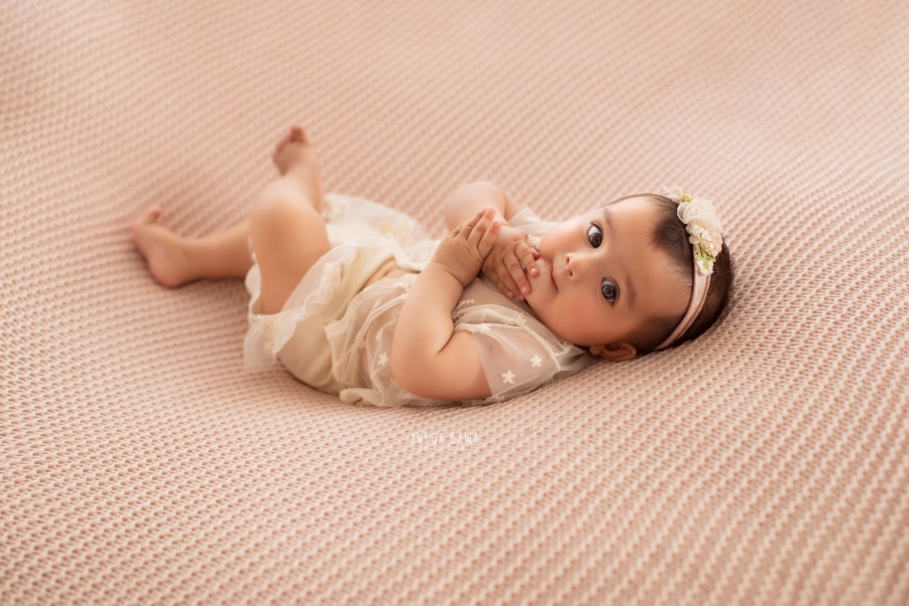 Captivating baby girl photoshoot by Anega Bawa, the esteemed baby photographer in Delhi, Gurgaon, and Noida, featuring the adorable 6, 5, 4, 3-month-old in a lying down pose, adorned with a cute headband against a serene beige backdrop.