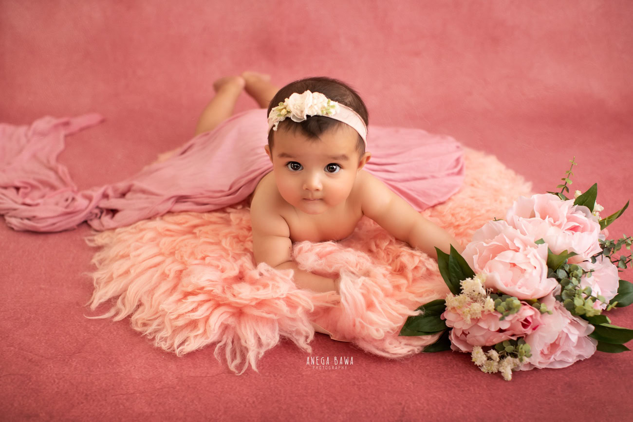 Captivating baby girl photoshoot by Anega Bawa, the esteemed baby photographer in Delhi, Gurgaon, and Noida, featuring the charming 6, 5, 4, 3-month-old adorned with a cute headband, wrapped in a pink wrap, on a lovely peach rug against a soft pink backdrop.