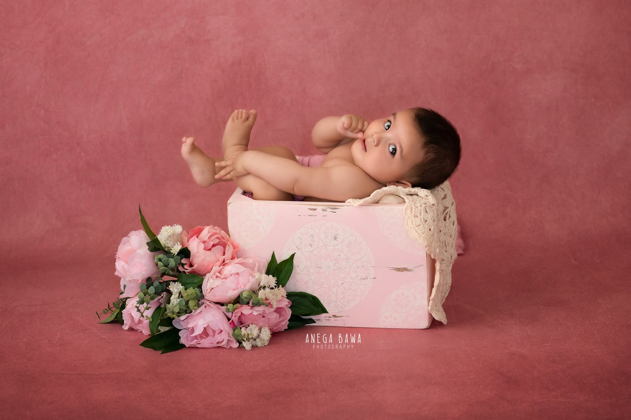 Captivating baby girl photoshoot by Anega Bawa, the esteemed baby photographer in Delhi, Gurgaon, and Noida, featuring the adorable 6, 5, 4, 3-month-old surrounded by flowers in a cute basket against a charming pink backdrop.