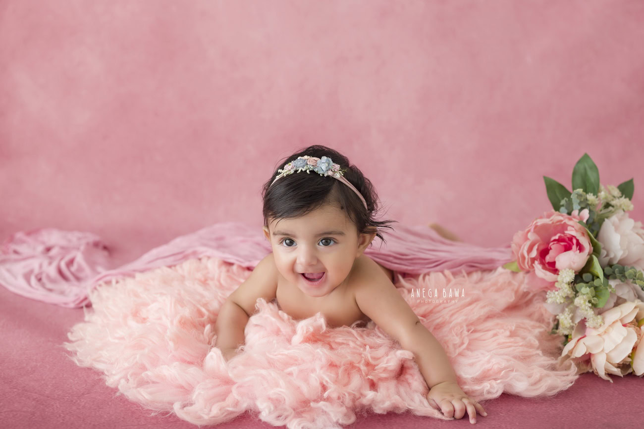 Baby girl photoshoot featuring a 3, 4, 5, or 6-month-old on a pink backdrop, wearing a cute headband and striking a smiling pose. Contact Anega Bawa for baby photoshoots in Delhi, Gurgaon, or Noida.